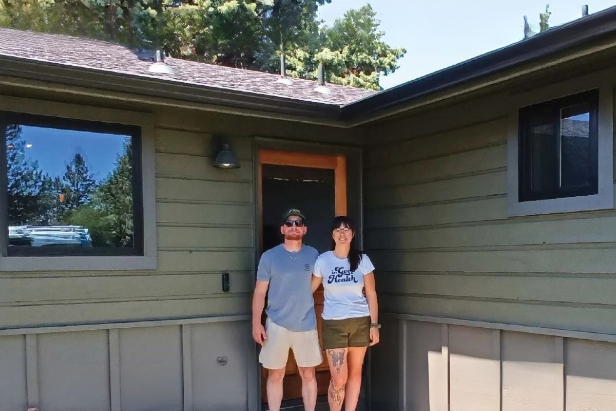 Two people stand in front of their front door linking arms.