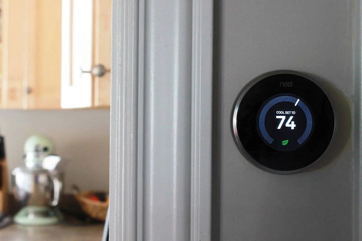 Nest thermostat mounted on a gray wall, with a modern kitchen visible in the background, showing smart home temperature control in a home setting.