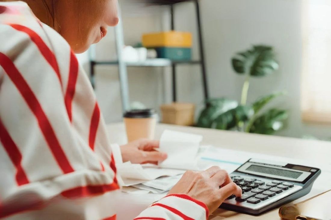 A person types on their calculator at a table with coffee. They are wearing a white shirt with red stripes.