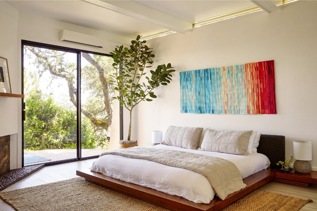 Ductless heat pump mounted on the wall of a bright white bedroom with a colorful painting, sliding glass door, and view of green trees outside.