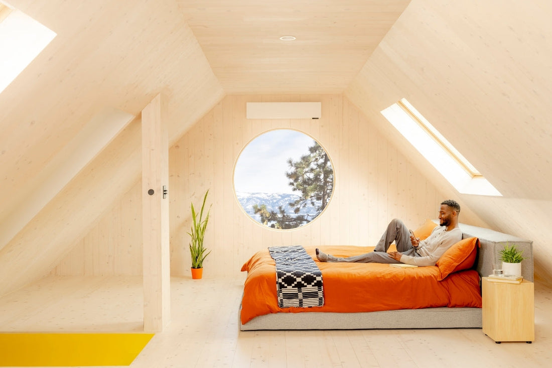 A person relaxes on their bed in a finished attic with a ductless heat pump mounted above a circular window.