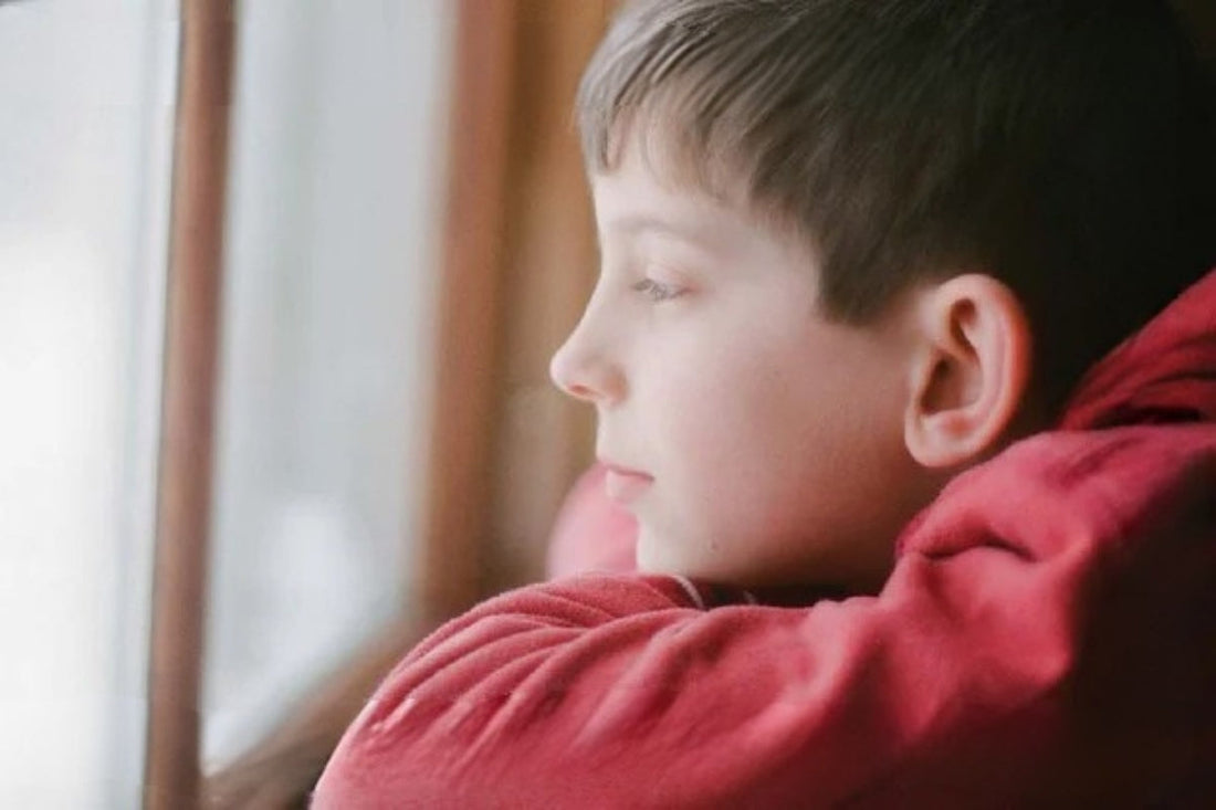 A child in a red sweatshirt rests his head on his hands and looks outside.