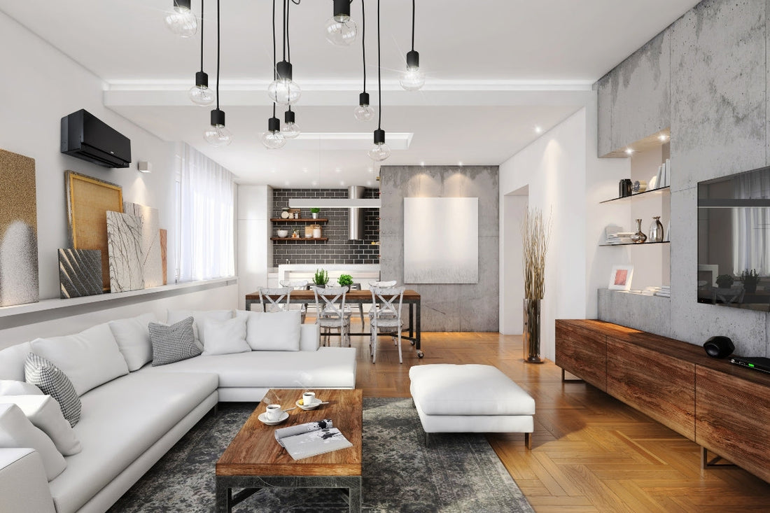 A homes modern living room with exposed light bulbs hanging down from the ceiling, a white sectional couch, wooden coffee tables and cabinets, and white and marbled walls.