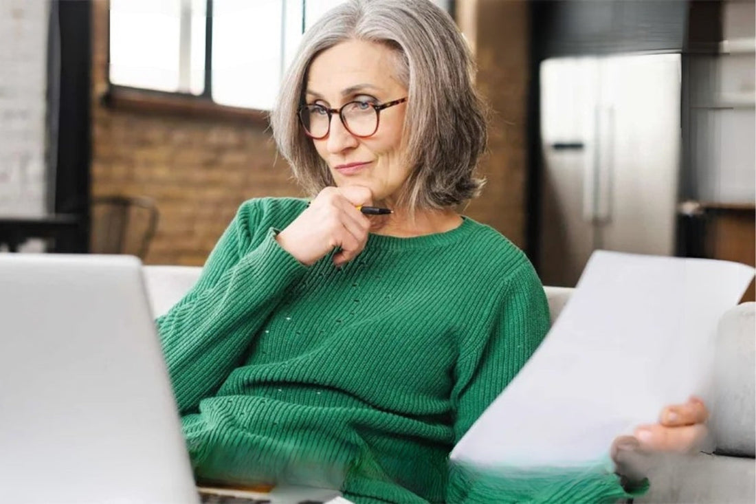 A person holds their hand to their chin and a paper in their other hand as they look at their laptop.