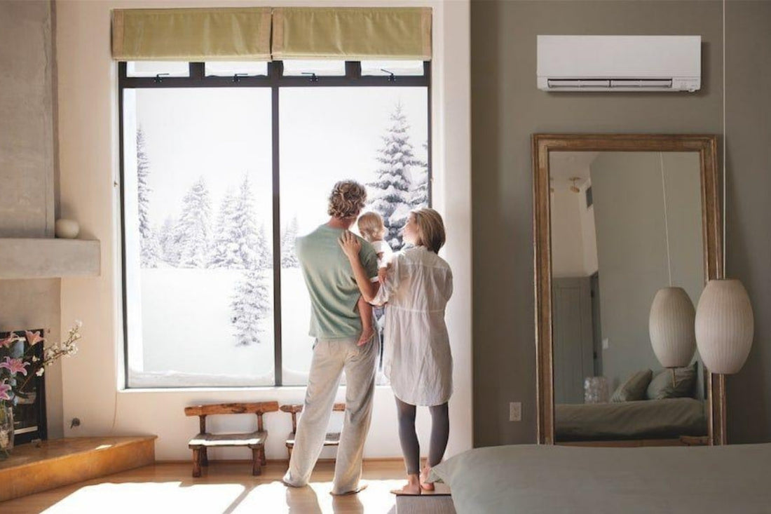 Two adults stand in their living room with one of them holding a child. They look outdoors at the snow.