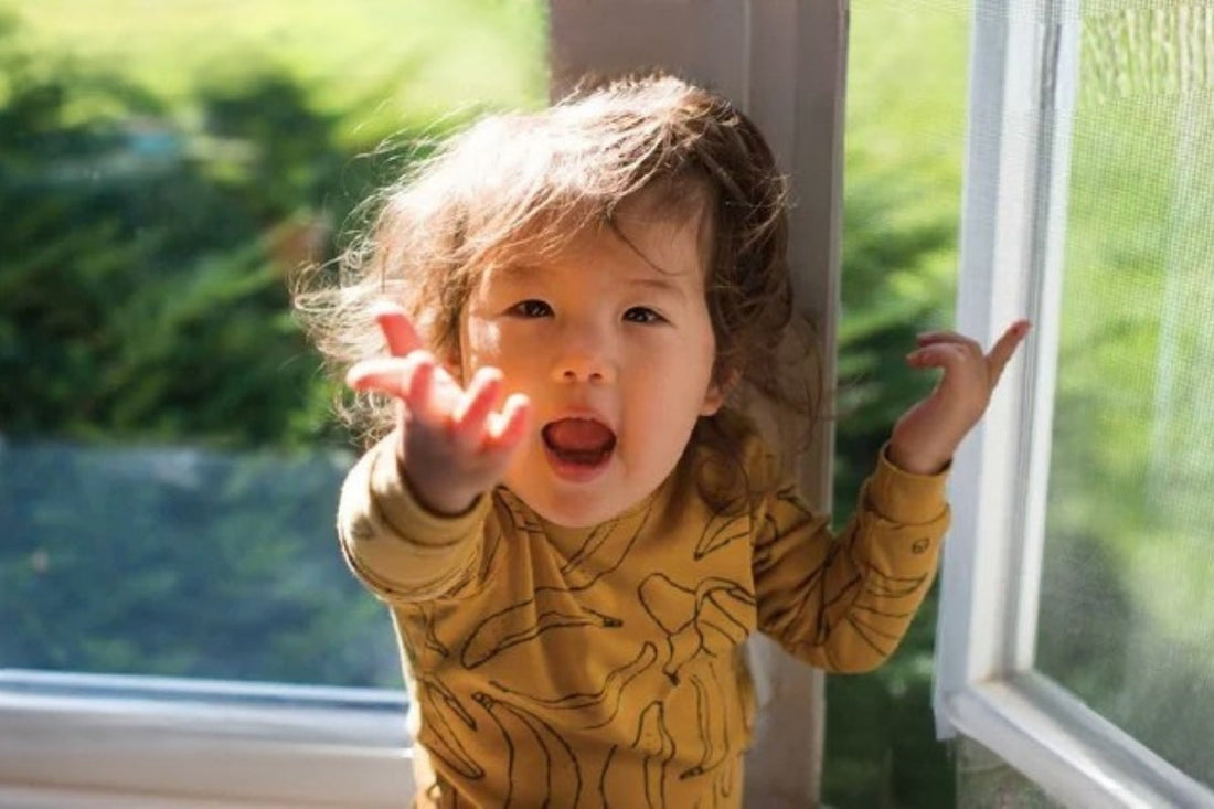 A child stands in front of a window that is ajar with their hands in the air.